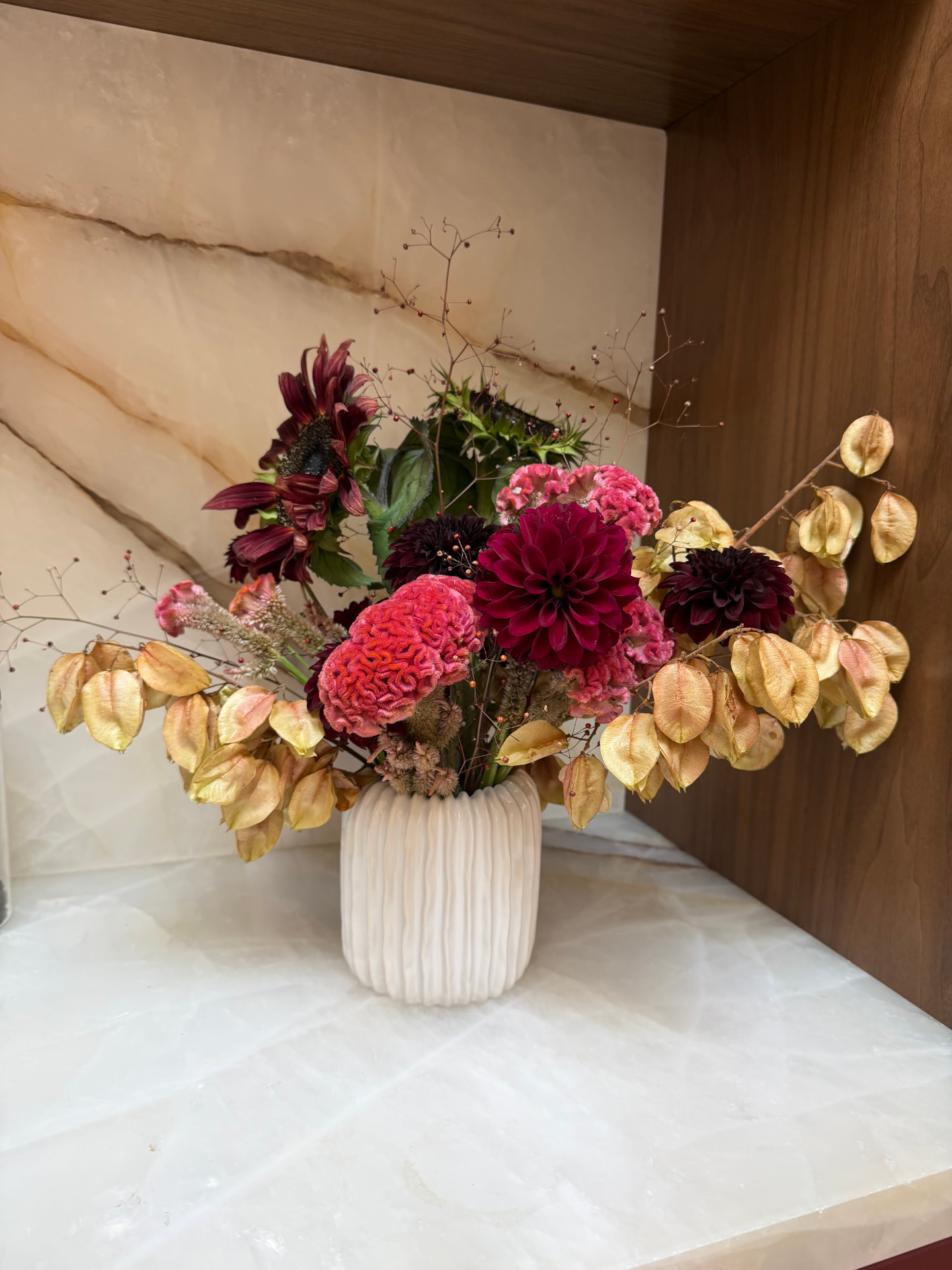 Burgundy dahlias, coral celosia and golden honesty pods in a fluted vase on an onyx shelf
