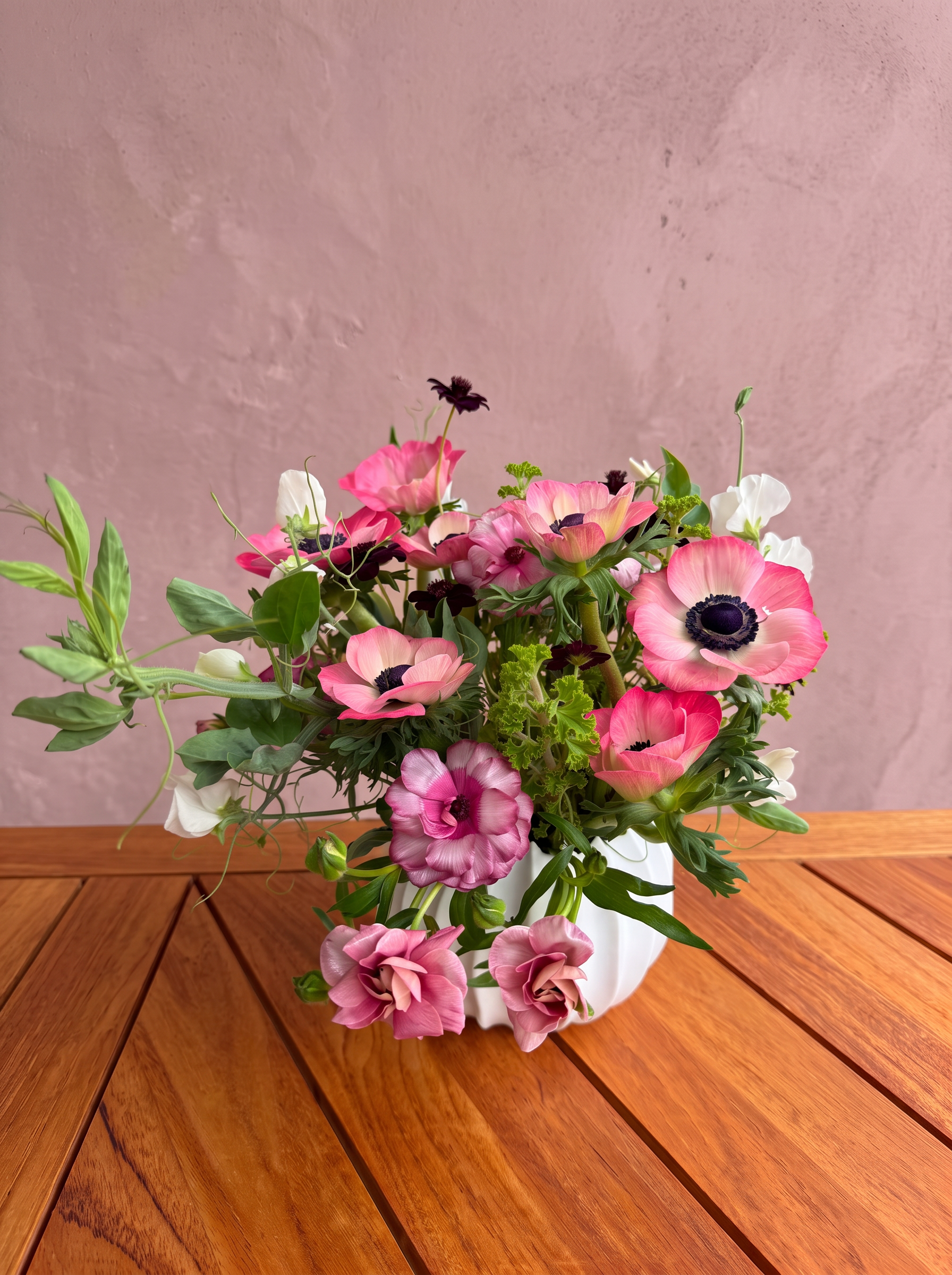 Pink anemones and trailing greenery in a white vase against dusty rose plaster