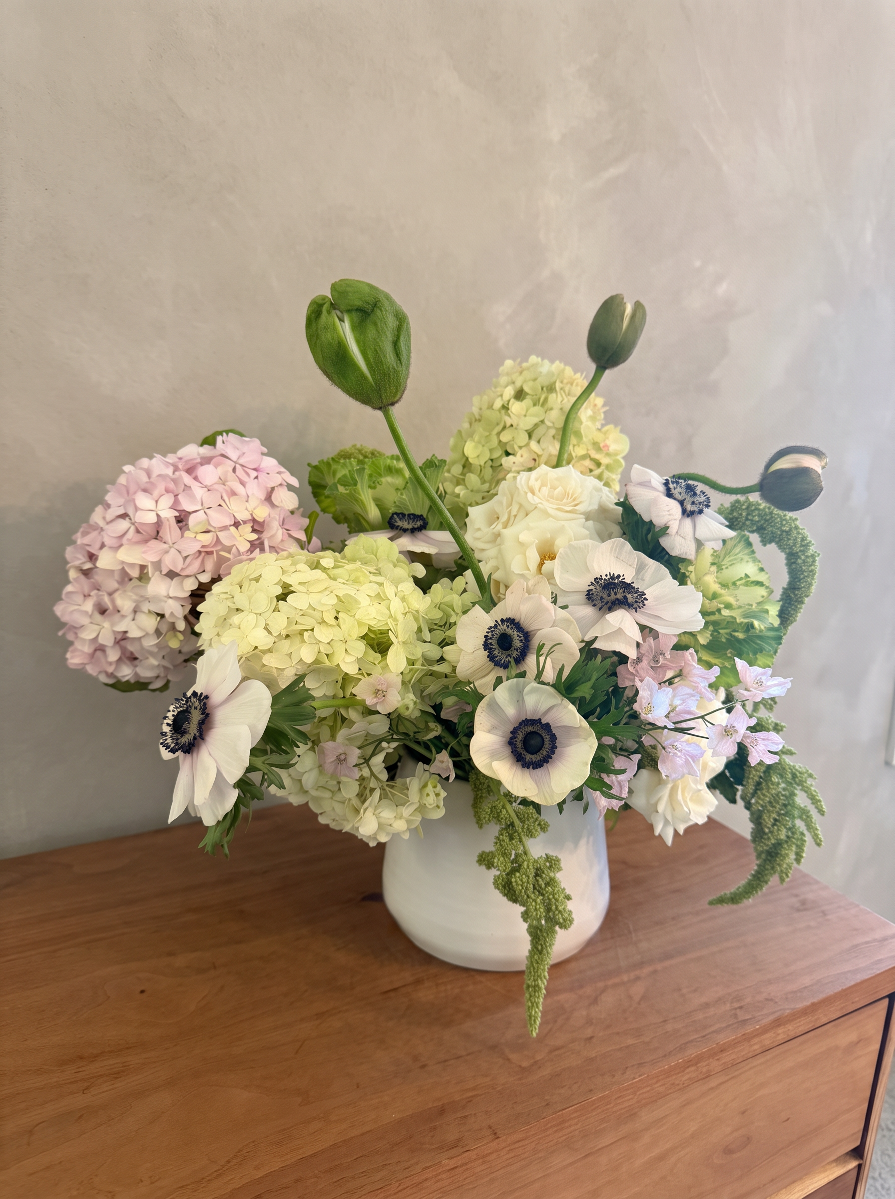 White anemones, pink hydrangea and trailing amaranth in a white vase on a walnut dresser