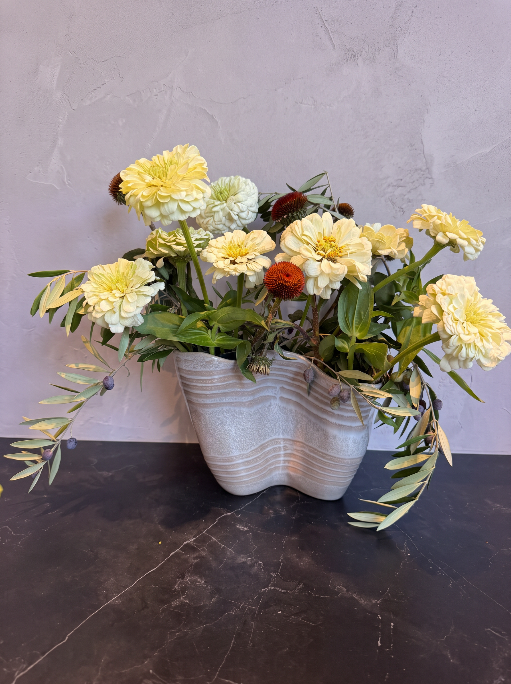 Cream zinnias, coneflowers and olive branches in a wavy vase against lavender-gray plaster